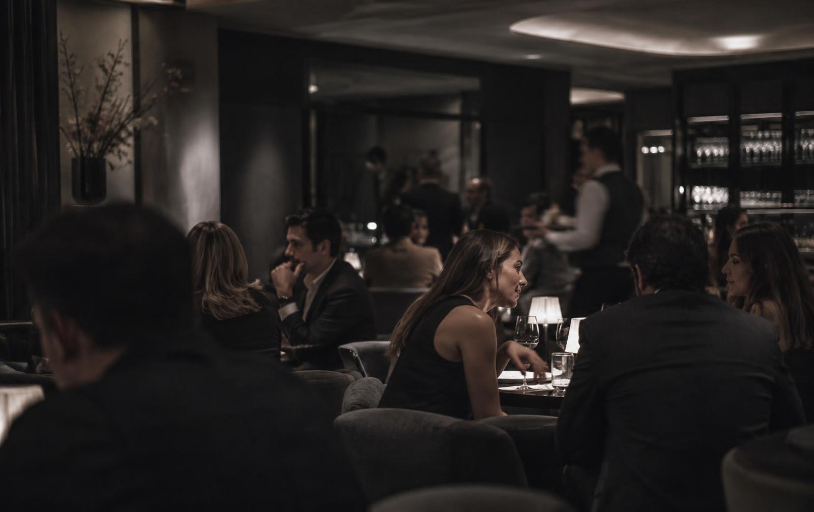 A crowded London bar at night, guests in conversation seen from across the room